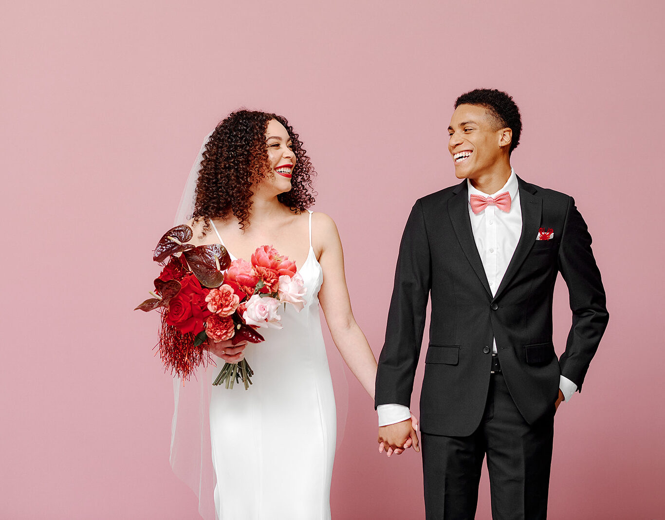 Couple laughing in front of a pink wall bride holding red bouquet groom wearing black tux with pink bow tie effortless-cool-affordable-luxury-wedding-gown-canadian-fashionable-bride-minimalist-wedding-dress-fun-minimal-bridal-wear